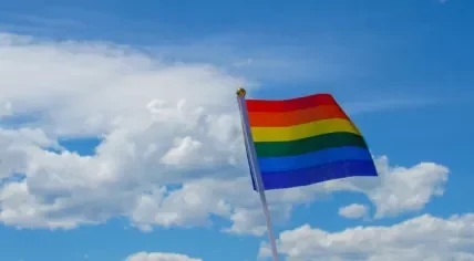 Rainbow pride flag against blue sky with clouds.